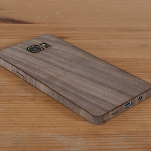 May include: A brown wood phone case with a textured wood grain pattern. The case has a camera cutout and a cutout for the charging port.
