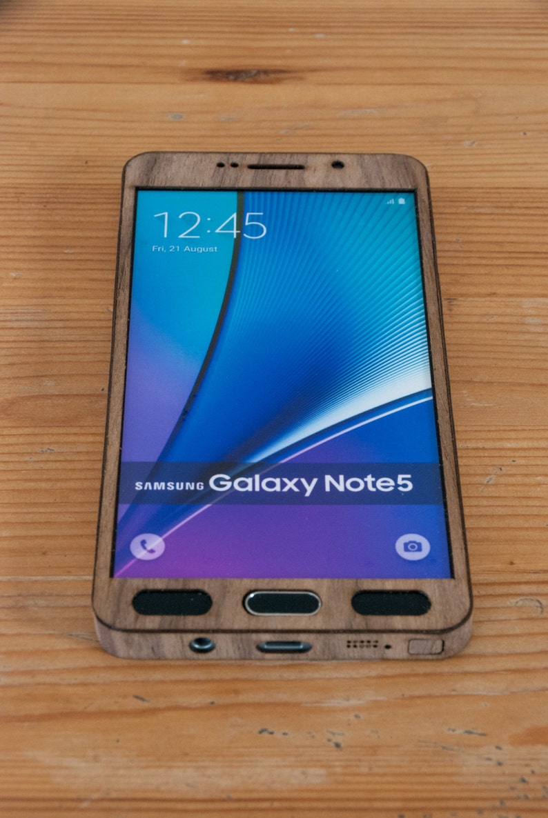 May include: A wooden phone case for a Samsung Galaxy Note 5. The case is made of dark wood and has a natural wood grain. The phone screen shows the time as 12:45 and the date as Fri, 21 August.