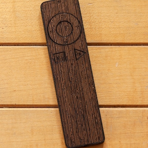 May include: A dark brown wooden remote control with a textured surface, featuring a circular navigation button and other control symbols. The remote is rectangular and lies on a light brown wooden surface.