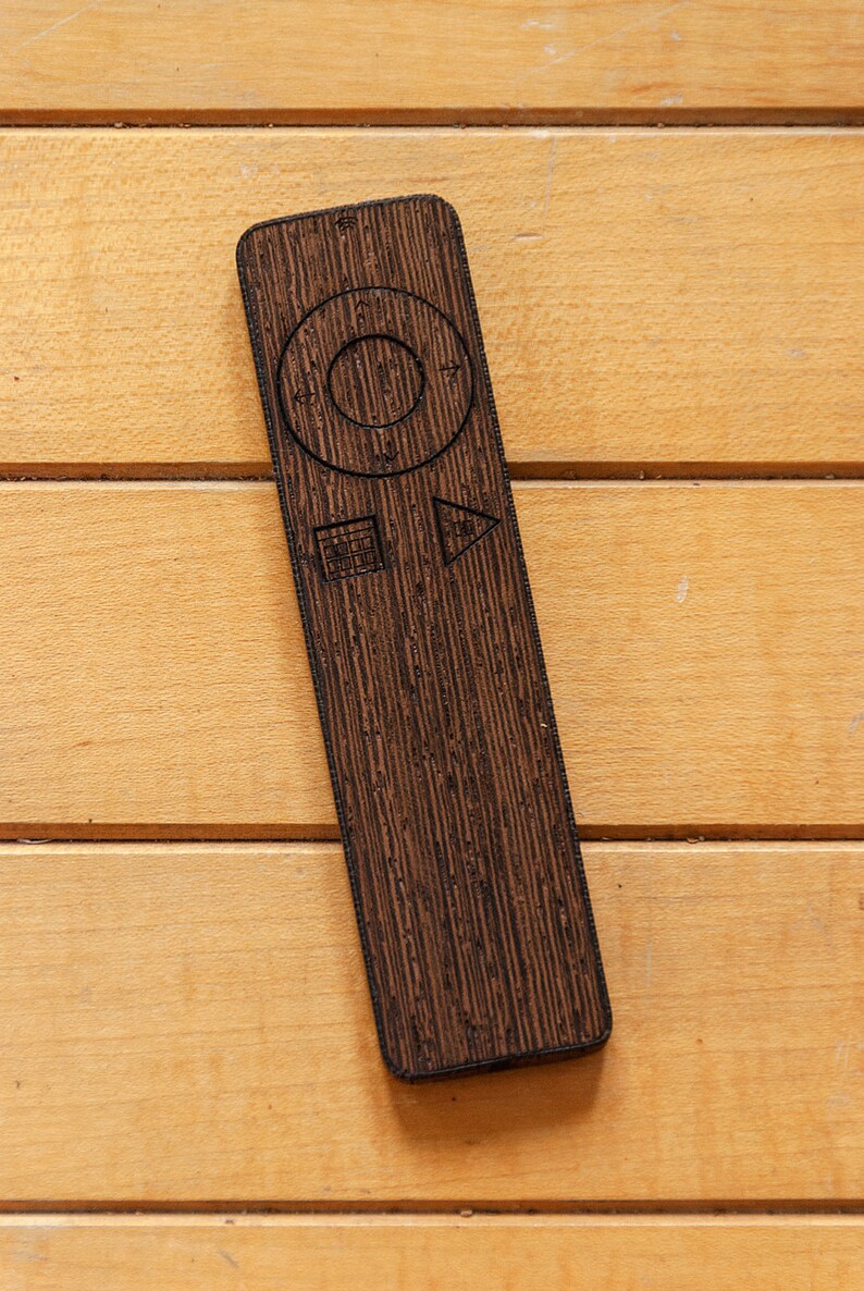 May include: A dark brown wooden remote control with a textured surface, featuring a circular navigation button and other control symbols. The remote is rectangular and lies on a light brown wooden surface.