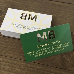 May include: Two business cards with the initials "MB" in gold foil lettering. The top card is white with a yellow circle pattern and the text "www.MBEmeraldEvents.com Santa Monica, CA". The bottom card is green with the text "Emerald Events Morgan Christiansen & Bergen Mesko 614-746-8003 / 917-992-0468 MB@MBEmeraldEvents.com".