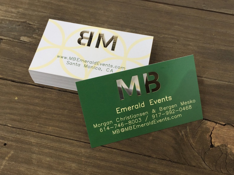 May include: Two business cards with the initials "MB" in gold foil lettering. The top card is white with a yellow circle pattern and the text "www.MBEmeraldEvents.com Santa Monica, CA". The bottom card is green with the text "Emerald Events Morgan Christiansen & Bergen Mesko 614-746-8003 / 917-992-0468 MB@MBEmeraldEvents.com".