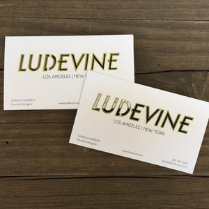 May include: Two white business cards with gold lettering. The text on the cards reads "LUDEVINE" with the locations "LOS ANGELES | NEW YORK" and "LOS ANGELES | NEW" printed below the name. The name of the founder and designer, "SHAHLA KAREEM", is printed on both cards along with the website address "www.ludevine.com". The phone number "350 741 7505" is printed on the bottom of one of the cards.