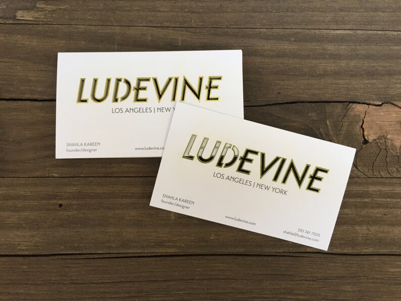 May include: Two white business cards with gold lettering. The text on the cards reads "LUDEVINE" with the locations "LOS ANGELES | NEW YORK" and "LOS ANGELES | NEW" printed below the name. The name of the founder and designer, "SHAHLA KAREEM", is printed on both cards along with the website address "www.ludevine.com". The phone number "350 741 7505" is printed on the bottom of one of the cards.
