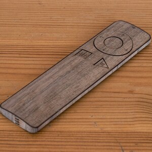 May include: A wooden remote control with etched symbols and a circular navigation pad. The remote is rectangular with rounded edges and a wood grain finish. It rests on a wooden surface.