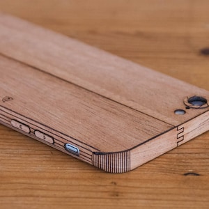 Puede incluir: Una funda de teléfono de madera con un acabado de madera natural. La funda tiene un borde elevado alrededor del objetivo de la cámara y un recorte para los botones de volumen. La funda está grabada con el texto "GRAINOR®".