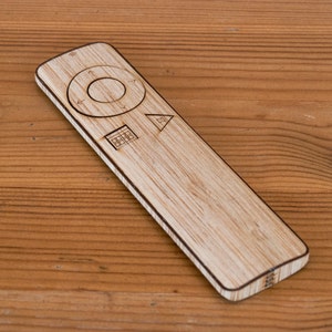 May include: A wooden remote control with a rectangular shape and rounded edges. The remote has a circular control pad, directional arrows, and other function symbols. The remote is made of light-colored wood and sits on a wooden surface.
