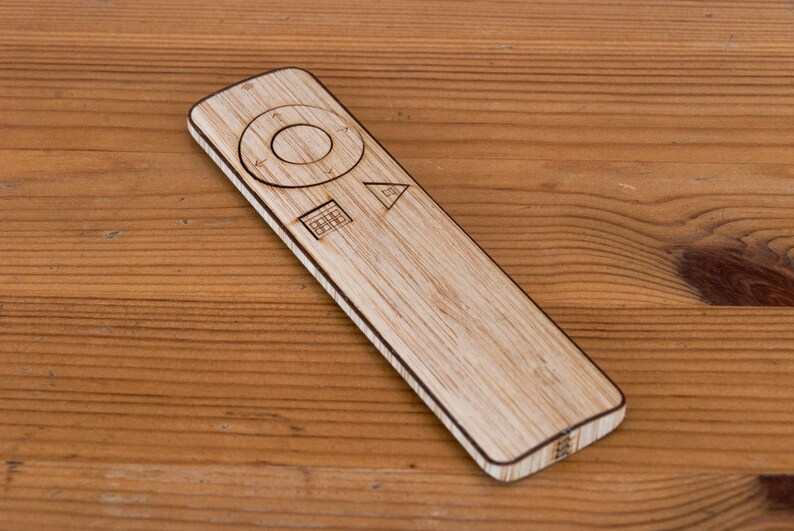 May include: A wooden remote control with a rectangular shape and rounded edges. The remote has a circular control pad, directional arrows, and other function symbols. The remote is made of light-colored wood and sits on a wooden surface.
