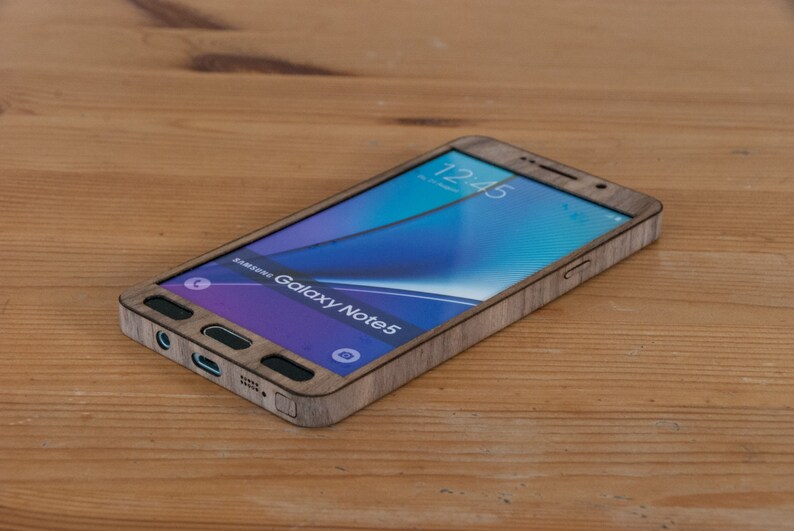 May include: A wooden phone case for a Samsung Galaxy Note 5. The case is made of real wood and has a natural wood grain finish. The phone is visible through the case, showing the time on the screen as 12:45.