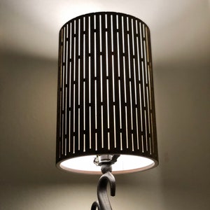 May include: A black lamp with a cylindrical lampshade made of wood with a slatted design. The lampshade is lit and the light shines through the slats.