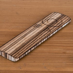 Puede incluir: Un mando a distancia de madera con un botón circular y un patrón a rayas. El mando está hecho de madera clara y oscura.