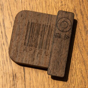 May include: A dark brown wooden device with a rectangular section and a rounded section. The rectangular section has a barcode and the word "APFEL" etched into it. The rounded section has control symbols etched into it.