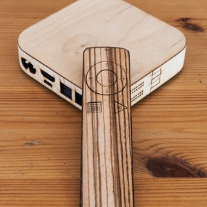 Puede incluir: Un conjunto de dispositivo multimedia y mando a distancia de madera. El dispositivo multimedia es un cuadrado de color claro con bordes redondeados. El mando es rectangular con símbolos de control. Ambos son de madera con un patrón de vetas rayadas.