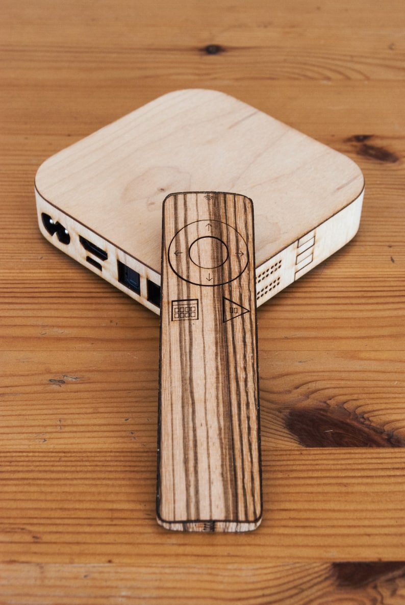 May include: A wooden media device and remote control set. The media device is a light-colored square with rounded edges. The remote is rectangular with control symbols. Both are made of wood with a striped grain pattern.