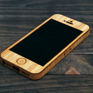 May include: A wooden phone case for an iPhone. The case is made of light brown wood and has a black screen. The case has a cutout for the camera and a small hole for the speaker.