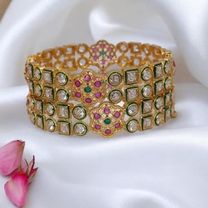 May include: Three gold bangles with intricate designs featuring white, pink, and green stones. The bangles are stacked on top of each other.