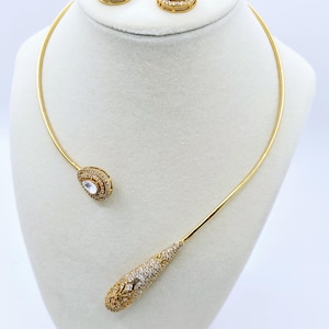 May include: A gold choker necklace with a teardrop-shaped pendant and a pair of matching earrings. The necklace and earrings are adorned with clear cubic zirconia stones.