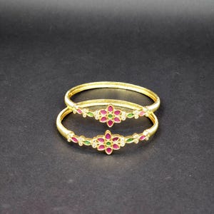 May include: Two gold bangles with intricate designs featuring pink and green gemstones. The bangles are stacked on top of each other.