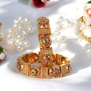 May include: A pair of gold bangles with intricate designs featuring red and green gemstones. The bangles are adorned with small, sparkling stones.