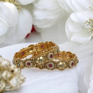May include: Two gold bangles with intricate floral designs and pink and white gemstones. The bangles are stacked on top of each other.