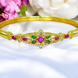 May include: A gold bangle bracelet with a decorative design featuring pink, green, and white gemstones.