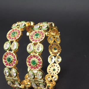 May include: A pair of gold bangles with intricate designs featuring white, pink, and green gemstones. The bangles are stacked on top of each other.