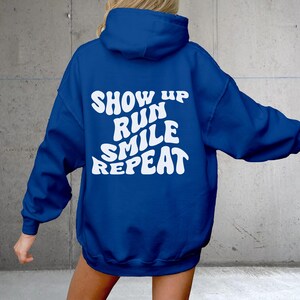 Puede incluir: Una sudadera con capucha azul real con el texto "SHOW UP RUN SMILE REPEAT" en blanco.
