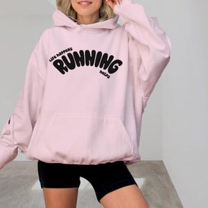 Puede incluir: Sudadera rosa de gran tamaño con capucha y un gráfico negro que dice "Life happens running helps".