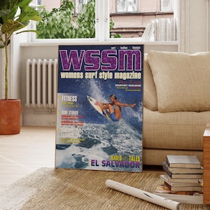 May include: A framed magazine cover for WSSM, Women's Surf Style Magazine, featuring a surfer riding a wave. The cover has a purple and white logo and the text "EL SALVADOR". The magazine is displayed on a rug near a window.