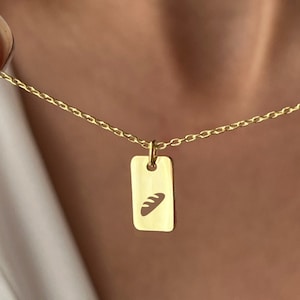 Può includere: Una collana color oro con un ciondolo rettangolare con un disegno di pane. Il ciondolo pende da una delicata catena. La collana è mostrata su una camicia bianca.