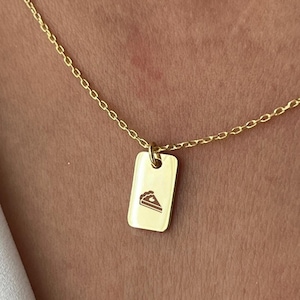 Puede incluir: Un collar de cadena dorada con un pequeño colgante rectangular con un diseño de rebanada de pastel.