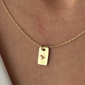 Könnte beinhalten: Eine goldene Kettenkette mit einem kleinen rechteckigen Anhänger, der eine Kolibri-Silhouette zeigt.