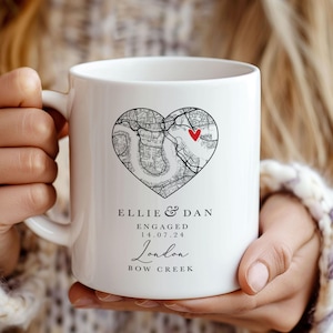 May include: White ceramic mug featuring a black and white map of London in the shape of a heart. The map has a red heart marking the location of Bow Creek. The text "Ellie & Dan Engaged 14.07.24 London Bow Creek" is printed below the map.
