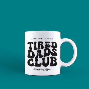 Puede incluir: Taza de cer&aacute;mica blanca con un asa grande. La taza presenta el texto "PROUD MEMBER OF THE TIRED DADS CLUB" en negro, en negrita, con una fuente de estilo retro. Debajo del texto est&aacute; el hashtag "#FueledbyCoffee". La taza est&aacute; sobre un fondo turquesa.