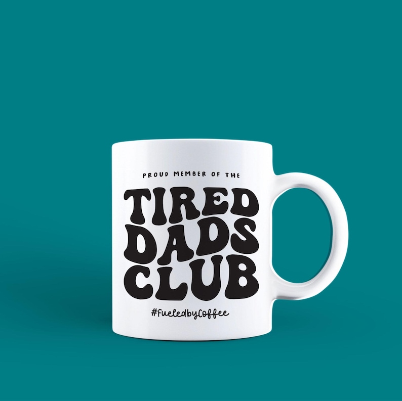 Puede incluir: Taza de cer&aacute;mica blanca con un asa grande. La taza presenta el texto "PROUD MEMBER OF THE TIRED DADS CLUB" en negro, en negrita, con una fuente de estilo retro. Debajo del texto est&aacute; el hashtag "#FueledbyCoffee". La taza est&aacute; sobre un fondo turquesa.