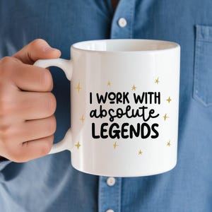 May include: White ceramic mug with a black inscription that reads "I work with absolute legends" and gold star accents.