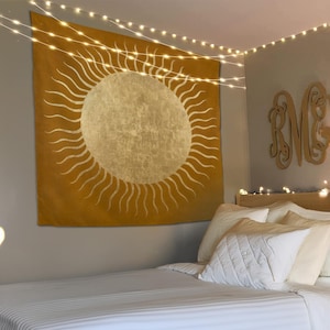 Könnte beinhalten: Senfgelbe Tapisserie mit einem großen Sonnendesign, das eine goldene Mitte und strahlende Linien aufweist. Die Tapisserie hängt über einem Bett mit weißer Bettwäsche und Kissen. Lichterketten sind an der Decke drapiert.