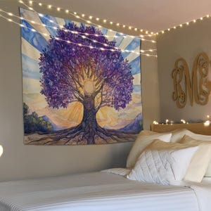 May include: A tapestry featuring a watercolor painting of a purple tree with a sun and rays of light behind it. The tree has intricate roots and a mandala design in the center of the trunk. The tapestry is hanging on a wall in a bedroom with a white bed and pillows.