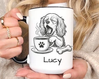 May include: White ceramic mug with a black rim and handle, featuring a black and white illustration of a yawning dog with a coffee cup and bone. The name "Lucy" is printed below the illustration.