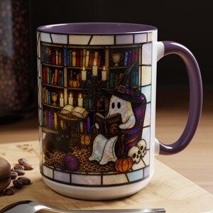 May include: A ceramic mug with a purple interior and handle, featuring a stained-glass style illustration of a ghost in a witch hat reading a book titled Dragons in a library, accompanied by a black cat, pumpkins, and skulls on a leopard print rug.
