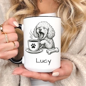 May include: White ceramic mug with a black rim and handle, featuring a black and white illustration of a yawning poodle with a coffee cup and bone. The name "Lucy" is printed below the illustration.
