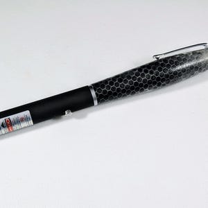 May include: A black laser pointer with a silver metallic tip and a silver pocket clip. The barrel features a black and grey hexagonal honeycomb pattern. A white warning label with red text is visible on the lower section of the device.