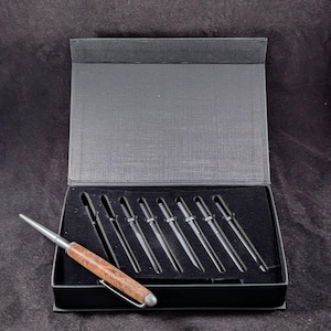 May include: A set of eight pens in a black presentation box. One pen is outside the box, showcasing a wood-grain barrel and silver accents. The other pens are black with silver tips. The box is open, revealing a black interior.