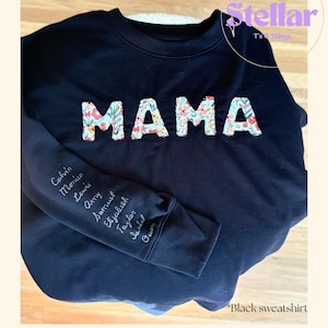 May include: A black crewneck sweatshirt featuring the word MAMA in large, floral-patterned fabric letters across the chest. The right sleeve is embroidered with a list of names including Calvin, Monica, Lewis, Amy, Samuel, Elizabeth, Taylor, Isabel, and Owen.