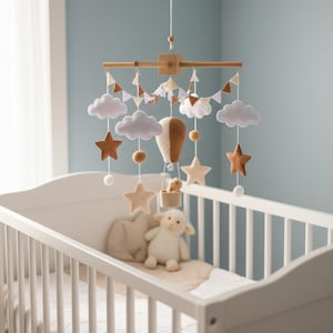 May include: A baby mobile made of wood, featuring felt clouds, stars, and a hot air balloon. It hangs above a white cot with a stuffed lamb and pillow. The colour scheme includes white, brown, and tan.