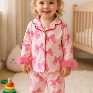 May include: A young child wearing a pink pyjama set patterned with bows and red trim. The pyjamas have feather trim on the cuffs and ankles. The child is smiling in a bright room.