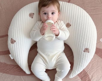 May include: A baby wearing a white onesie is lying on a cream-colored nursing pillow. The pillow has a crescent shape with a quilted texture and embroidered bear accents. The baby is holding a clear bottle.