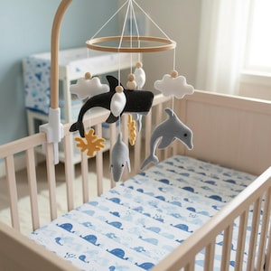 May include: A baby's cot with a wooden frame and a blue and white patterned sheet. A mobile hangs above the cot, featuring felt whales, dolphins, clouds, and coral. The mobile is suspended from a wooden arm attached to the cot.