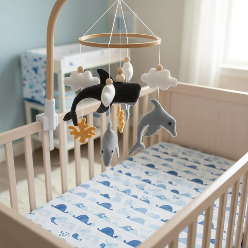 May include: A baby's cot with a wooden frame and a blue and white patterned sheet. A mobile hangs above the cot, featuring felt whales, dolphins, clouds, and coral. The mobile is suspended from a wooden arm attached to the cot.