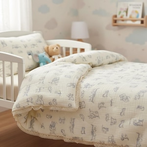 May include: A cream-coloured baby bedding set with a blue cartoon animal print. The set includes a duvet, pillow, and bumper, all with a delicate lace trim. The bedding is on a white cot.
